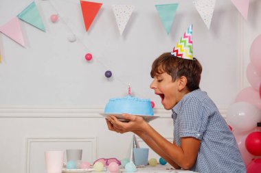Parti şapkalı neşeli genç bir çocuk elinde bir mumlu mavi bir pasta tutuyor. Ağzını kocaman açıyor. Canlı doğum günü süslemeleri arasında balonlar ve kiraz kuşu da var.