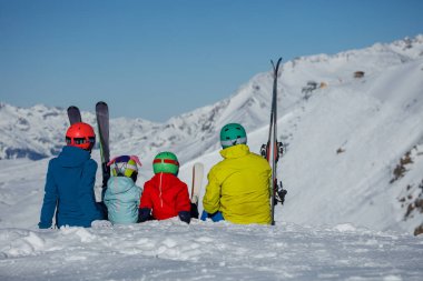 Kayak kıyafeti giymiş küçük çocukları olan dörtlü bir aile, karlı bir tepenin tepesinde oturmuş, görkemli, karla kaplı dağlara bakıyor.