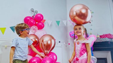 Şirin küçük çocuklar etrafları parlak pembe balonlarla çevrili bir partide eğleniyorlar. Kız renkli bir peri gibi giyinmiş. Arka planı süslüyor.