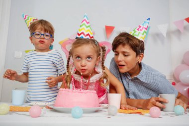 Pembe bir doğum günü pastası etrafında toplanan üç çocuk yanan mumları söndürmeye hazırlanıyor.