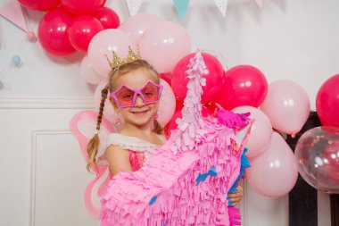 Prenses gibi giyinmiş genç bir kız elinde bir hediye olan tek boynuzlu at pinatasının yanında duruyor. Parlak pembe balonlar odayı süslüyor.