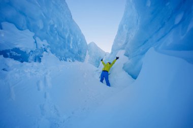 Karlı bir buz kanyonunda canlı sarı bir palto giymiş bir adam sevinçle ellerini kaldırır.