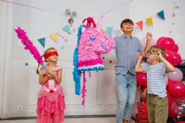 Renkli süslemelerle dolu bir odada, gözleri bağlı bir çocuk ve peri elbisesi iki hevesli erkek tarafından cesaretlendirilen tek boynuzlu at pinatasına bir sopa doğrultuyor.