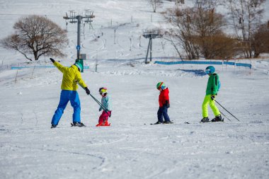 Yamaçlarda, canlı kış kıyafetleri içinde üç çocuk karda temel teknikleri öğrenirken bir kayak hocasının yolunu takip ediyor.