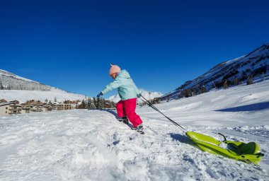 Pastel mavisi ceketli küçük bir kız tatil sırasında açık mavi gökyüzünün altında yeşil bir kızak çekiyor.