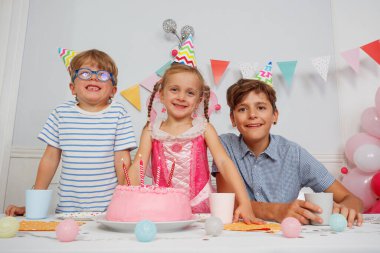 Bir grup mutlu çocuk, kafalarının üstünde mumlar ve parlak şapkalar olan pembe bir pastayla kutlama yapıyorlar.
