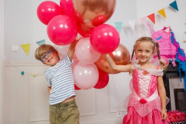 Parlak balonlarla dolu bir kutlama sahnesinde, mavi gözlüklü neşeli bir çocuk pembe peri gibi giyinmiş bir kızın yanında poz veriyor.