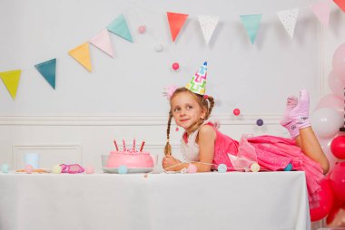 Kutlama için giyinmiş bir kız masaya uzanmış pembe pastanın yanında dilek diliyor, yukarıda doğum günü mumları ve neşeli afişlerle