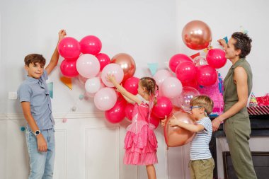Güzel bir aile doğum gününü pembe ve altın balonlarla kutluyor. Taburede dikilen süslü elbiseli küçük bir kız. Yanlarındaki masada renkli bir kağıda sarılmış hediyeler.