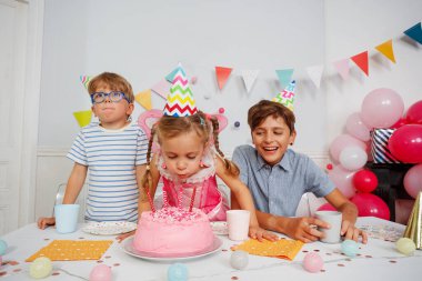 Bir grup çocuk, kız, renkli şapkalı iki oğlan pembe bir pasta etrafında, doğum günü mumlarını üfleyin.