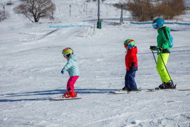 Karda, kask ve renkli kayak kıyafetleriyle donatılmış üç çocuk yokuş aşağı kayıyor.