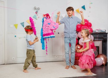 Gözleri bağlı genç bir çocuk renkli bir pinataya vurmaya hazırlanırken diğer çocuklar şenlikli bir partide hevesle izliyorlar.