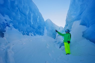 Bu sahnede, parlak yeşil kayak kıyafeti ve sarı pantolon giyen bir kayakçı elinde kayaklarla, kaskını takmış, görkemli buz duvarlarıyla çevrili duruyor.