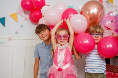 Neşeli bir sahnenin ortasında, üç çocuk parlak balonların ve çeşitli parti afişlerinin yanında gülüyor.