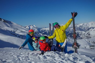 Çocuklu ebeveynler karlı bir dağda kayak yapmayı severler, açık mavi gökyüzü ve arka planda manzaralı zirveler.