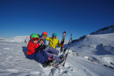 Dört kişilik bir grup, anne baba iki çocuk karlı bir yamaçta neşeli bir şekilde oturuyor, açık mavi gökyüzünün altında canlı kayak kıyafetleri giyiyorlar, arka planda dağlık bir manzara var.