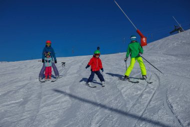 Bir kayakçı üçlüsü kış tatillerinde açık mavi bir gökyüzünün altında karlı bir yamaçta birlikte poz verir.