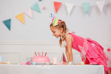 Aydınlık bir odada, parti şapkası takan bir çocuk ilginç bir şekilde masasında mumlar olan, canlı dekorasyonlarla çevrili pastaya bakar.