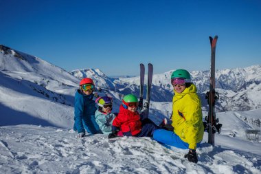 Güneşli bir kış gününde, canlı kayak kıyafetleri giymiş neşeli bir grup, karlı bir tepenin üzerinde, manzaralı dağ manzaralarıyla çevrili bir şekilde poz verirler.