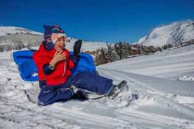 Kırmızı ceketli gülen çocuk kızaktan düştü. Karlı dağ tepesinde iyi eğlenceler.