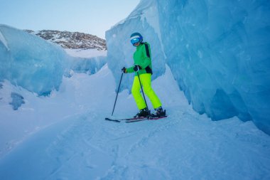 Genç çocuk kayaklarla poz veriyor, parlak kıyafetler giymiş, buz duvarlarının önünde duruyor ve parlak bir miğfer takıyor.