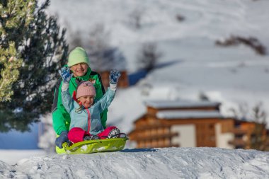 İki çocuk karla kaplı bir yamaçtan aşağı zum yapıyor. Arkalarında kışlık tepeler ve binalar var.