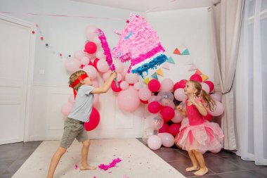 Bir parti sahnesinde, gözleri bağlı bir çocuk yanında neşeli bir kızla renkli bir pinataya yumruk atmak üzere.