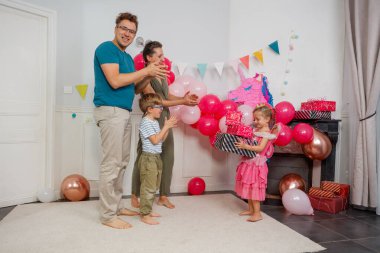 Pembe ve altın balonlardan oluşan şenlikli bir düzende, küçük bir kız elbise giyer, arkasında hediyelerle aile toplanır.