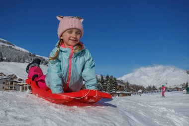 Canlı bir kış ortamında, örgülü saçlı bir çocuğun karlı bir yamaçtan sıcak ve komik bir şapkayla kaydığı görülüyor.