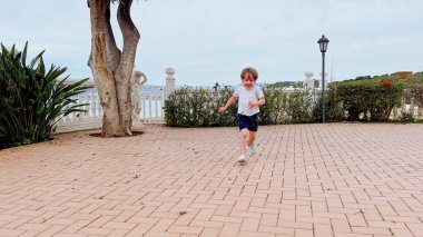 Bir çocuk neşeyle kaldırımlı bir avluda koşuyor, arka planda bir ağaç ve yeşillik bulutlu bir gökyüzünün altında