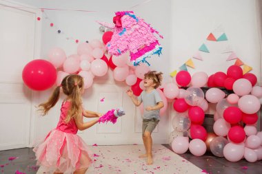Prenses gibi giyinmiş bir kız, gözleri bağlı, dekoru dolu bir odada oğlanın coşkulu desteğiyle tek boynuzlu at pinatasını kırmaya çalışıyor.