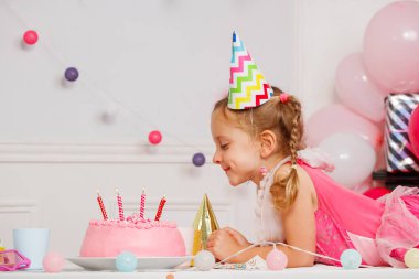 Küçük bir kız çok renkli bir parti şapkası takıyor. Kutlama balonları ve ışıklarının önünde mumlarla pastayı neşeyle izliyor.