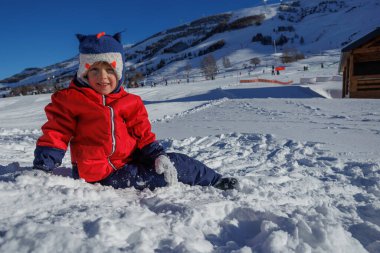 Kırmızı ceketli, mutlu bir genç kar üzerinde uzanmış, dağlarda güzel bir kış gününün tadını çıkarıyor.