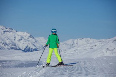 Neon yeşil ceketli genç bir kayakçı kış tatillerinde açık mavi gökyüzünün altında kayak yapıyor.