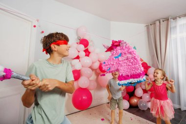 Etrafı parti süslemeleriyle çevrili, gözleri bağlı bir çocuk, bir kızın oturma odasındaki anın keyfini çıkarırken bir pinataya vurmak üzere.