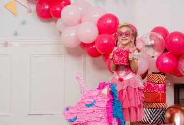 Prenses elbisesi giymiş bir çocuk elinde pembe balonlarla sarılı bir hediye taşıyan renkli bir tek boynuzlu atın yanında duruyor.