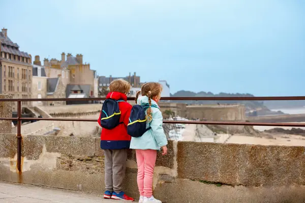 Sırt çantalı iki çocuk, Fransa 'nın Bretagne kentindeki taştan bir bariyerin arkasından Saint-Malo kıyı kasabasına bakıyor.