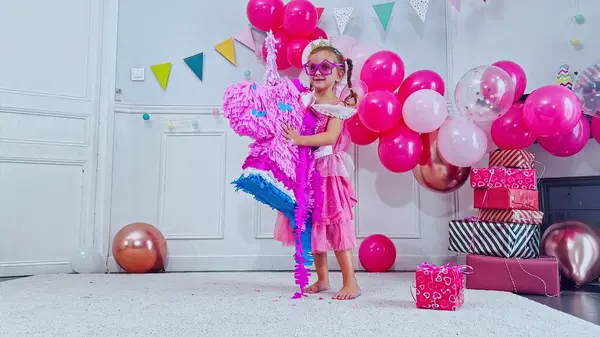 Çocuk mor gözlük takıyor, taç pembe bir tek boynuzlu at pinyatası taşıyor, çeşitli balonlar ve asılı bayraklarla parti benzeri bir ortamda.