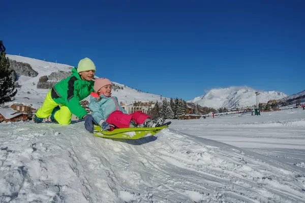 Nane paltolu çocuk kızağa binmiş küçük kız kardeşini dürtüklüyor, karlı tepelerden önce, kayak tatillerinde eğlence.