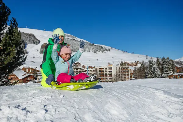 Neon yeşil ceketli bir çocuk, kış tatillerinde tatil köyü yakınlarındaki bir kızakta pembe pantolonlu bir arkadaşını itiyor.