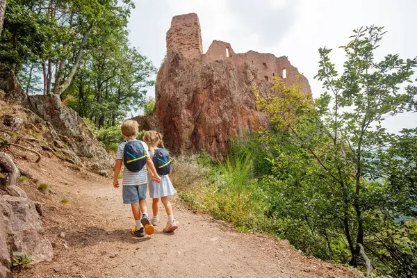 Sırt çantalı iki gülen çocuk, arka planda eski bir şato ve yemyeşil tepeler olan bir patikada duruyorlar.