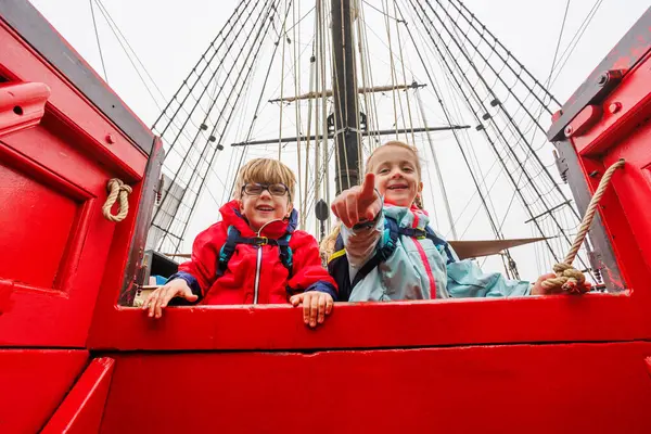 Heyecanla, grev ceketli iki çocuk kırmızı bir teknenin kenarından, gemi direğinin altından dışarı bakıyor.