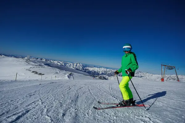 Gökyüzü açık, parlak yeşil kayak kıyafeti giyen genç bir kayakçı dağ eteklerinde karlı bir yokuş aşağı iniyor.