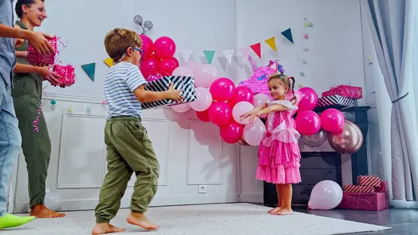 Pembe prenses kostümü giymiş bir çocuk balonlar ve renkli süslemelerle çevrili bir şenlik partisinde iki çocuğun hediyesini hevesle kabul eder.