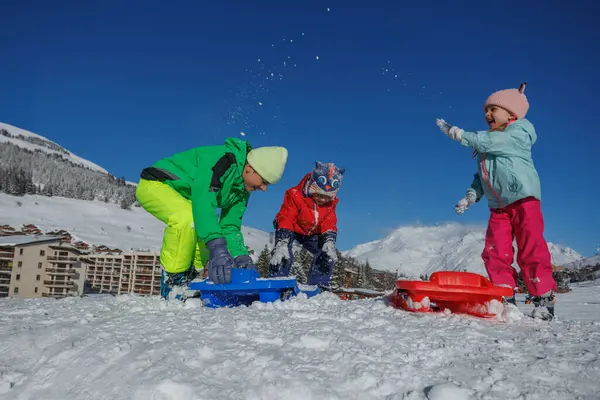 Kış kıyafetleri giymiş bir grup çocuk, karlı bir ortamda ve parlak mavi bir arka planda kar topu savaşında eğleniyor.