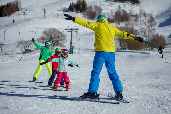Karlı bir dağdaki bir eğitmen üç çocuğa kayak teknikleri gösteriyor. Hepsi Alp Otel 'de göz alıcı kışlık giysiler giymiş.