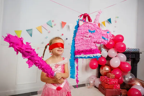 Etrafı şenlikli dekorlarla çevrili pembe kostümlü bir kız doğum günü partisinde tek boynuzlu at pinatasına vurmaya çalışıyor.