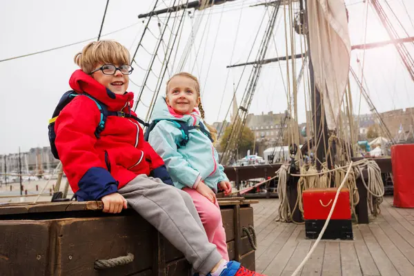 Parlak ceketler giyen iki çocuk, bir yelkenli teknesinde ahşap bir sandıkta oturmuş, ziyaretlerinin tadını çıkarıyorlar.