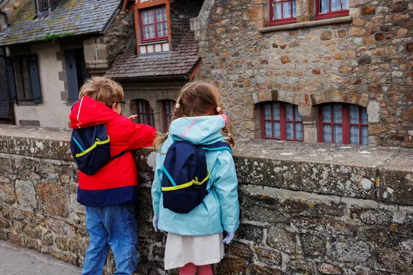 İki çocuk, geleneksel bir eve ve yapıyı işaret eden bir parmağa dikkatlerini vererek bir duvarın yanında duruyorlar.