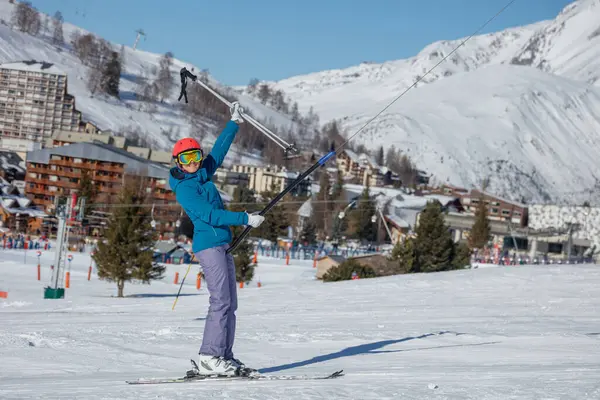Canlı bir sahne, kar kaplı bir yamaçta mavi ceket ve kırmızı kask giyen bir kayakçıyı ve uzaktaki geleneksel kayak kulübelerini gösteriyor.